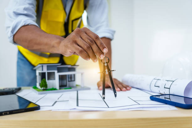 Architect works at desk with house model toy and A3 paper blueprints, construction real estate law, zoning, permits, and resolving property disputes through arbitration and litigation.