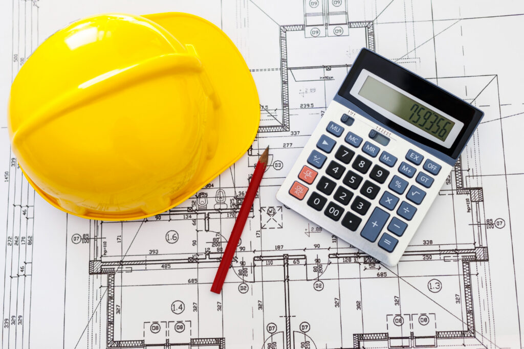 Blueprint with a calculator, tape measure, notebook, and construction cost estimate spread across a table.